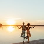 Two women practicing yoga by a tranquil lake during sunset, perfect blend of fitness and nature.