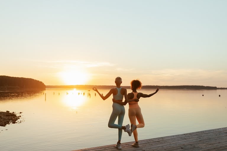 Two women practicing yoga by a tranquil lake during sunset, perfect blend of fitness and nature.