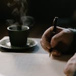 A person writing in a journal or a habit tracker with a cup of coffee beside them