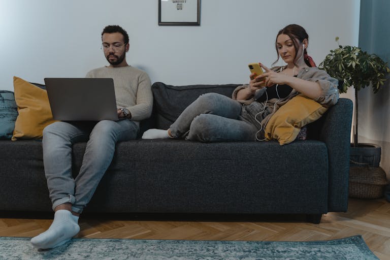 A person using a laptop while sitting on a couch with their phone nearby, depicting the balancing act between work and personal life.