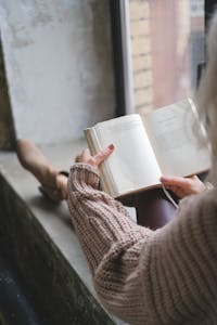 A person placing their phone in a drawer while reading a book beside a window.