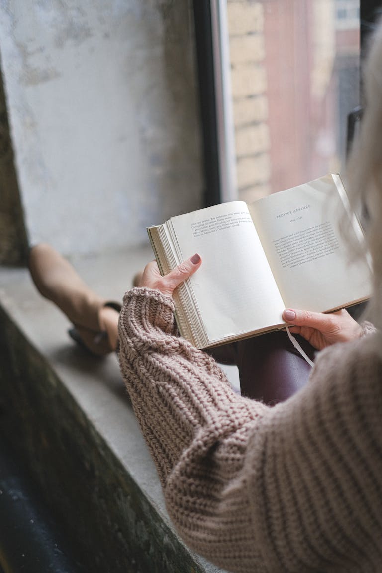 A person placing their phone in a drawer while reading a book beside a window.