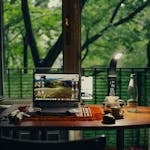 A home workspace with a coffee mug, notebook, and a laptop, surrounded by greenery and natural light.