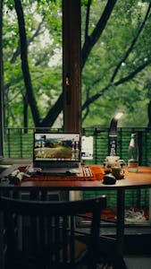 A home workspace with a coffee mug, notebook, and a laptop, surrounded by greenery and natural light.