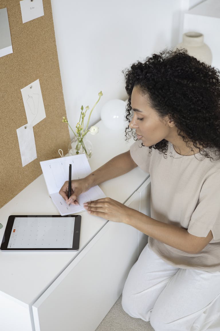 A person writing goals on a notepad, with a vision board and calendar in the background.