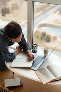 A person reading a book at a desk, surrounded by notes, coffee, and a laptop showing online courses.