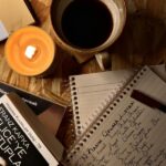A hand writing in a journal beside a candle and coffee mug.