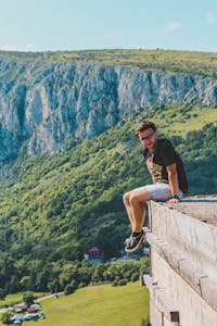 Man enjoys scenic mountain view sitting on cliff, capturing adventure and freedom.