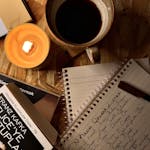 A hand writing in a journal beside a candle and coffee mug.