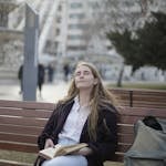 A serene individual sitting peacefully on a park bench, surrounded by nature, holding a cup of tea while traffic blurs in the background.