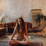 Woman meditating on a yoga mat with a laptop and mug in a stylish living room.